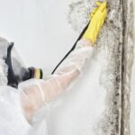 A professional wearing protective gear performing mold removal on a wall, highlighting the process of comprehensive mold remediation.