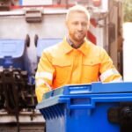 A waste removal worker in an orange uniform handles a blue trash bin near a waste truck, ensuring efficient waste management.