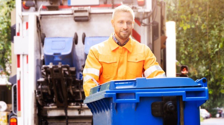 A waste removal worker in an orange uniform handles a blue trash bin near a waste truck, ensuring efficient waste management.