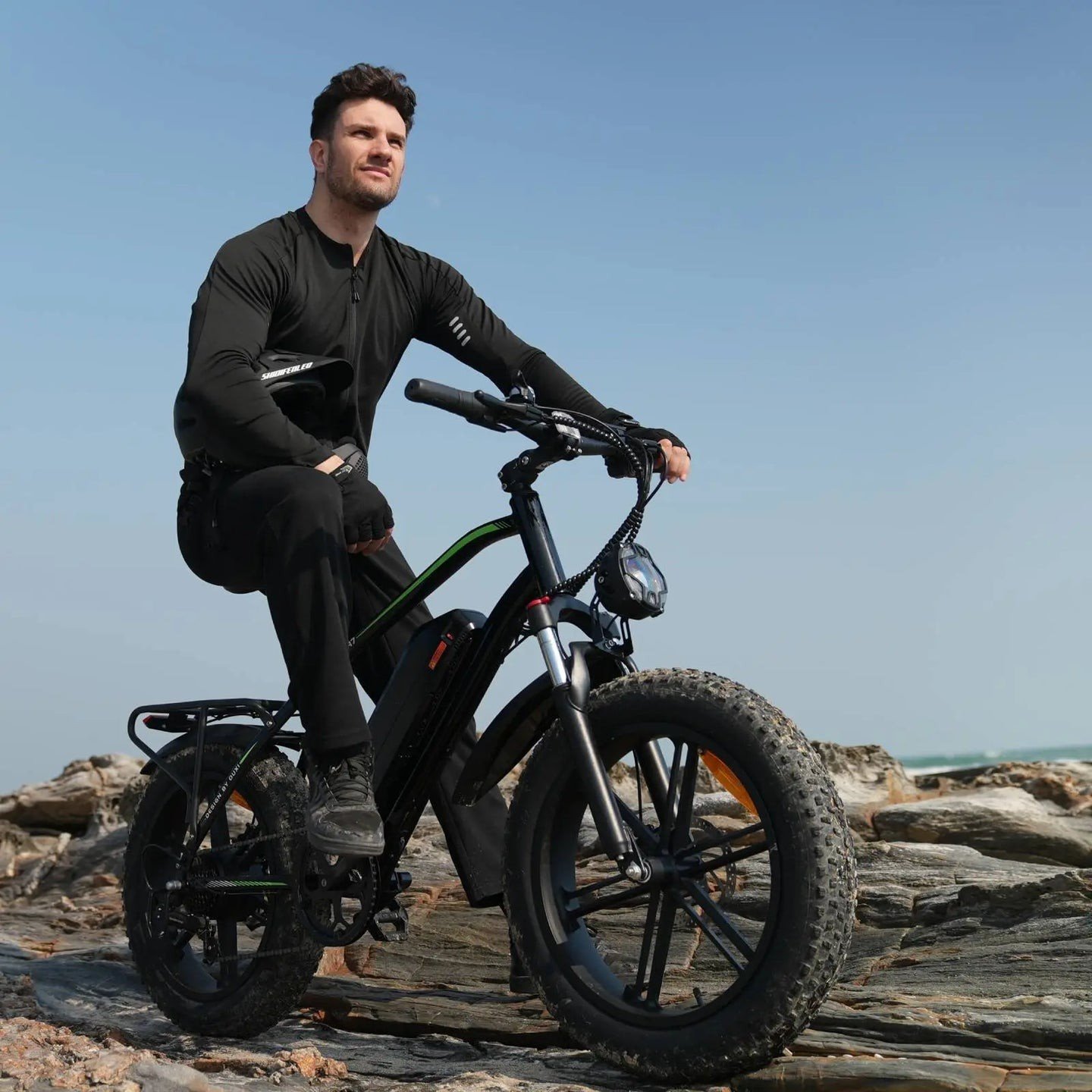 Man in Fat Tire Electric Bike sits confidently on a rugged e-bike atop rocky terrain under a clear blue sky, exuding adventure and determination.