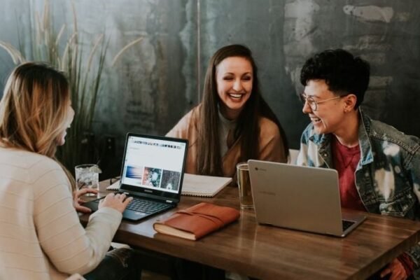Three people sit at a wooden table with laptops, smiling and engaged in conversation. The scene conveys a collaborative and positive atmosphere.