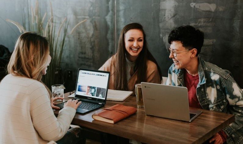 Three people sit at a wooden table with laptops, smiling and engaged in conversation. The scene conveys a collaborative and positive atmosphere.