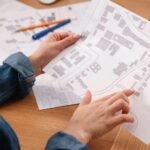 A land surveying company professional analyzes detailed maps, pointing to key areas for a property survey, with a laptop in the background.