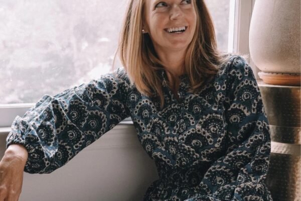 Smiling woman wearing a blue block print dress with intricate patterns, sitting by the window, showcasing comfort and style in handcrafted fashion