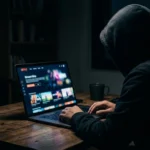 A hooded figure with hands on a laptop keyboard in a dark room. The screen displays a colorful media interface. A coffee cup sits on the wooden desk, and city lights are visible